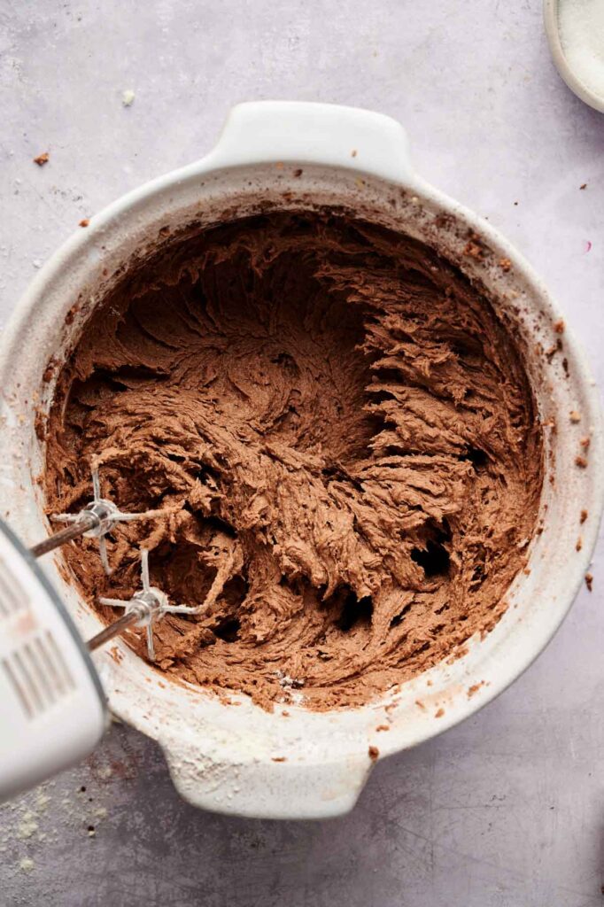 Electric hand mixer with beaters in a white bowl filled with whipped chocolate cake batter&mdash;perfect for making Air Fryer Cream Cheese Brownies&mdash;viewed from above on a light-colored surface.