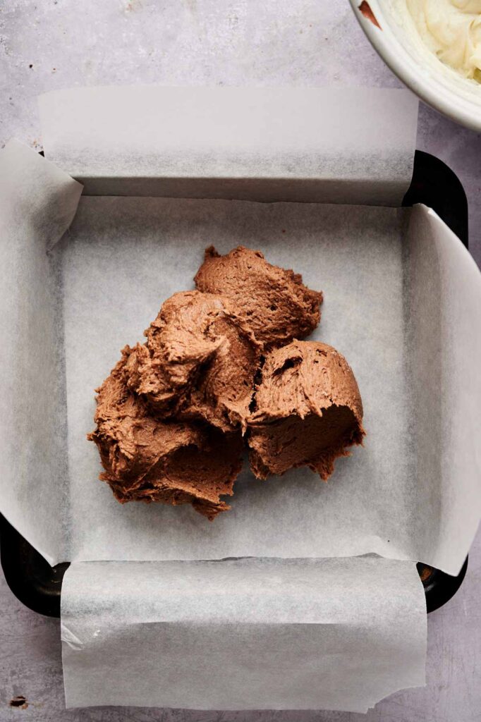 A square baking pan lined with parchment paper holds several scoops of chocolate batter, ready to be spread for delicious Air Fryer Cream Cheese Brownies.