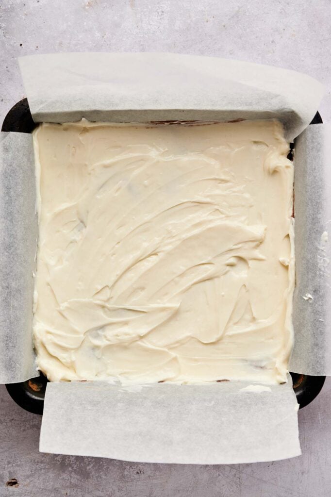 A baking pan lined with parchment paper, filled with a smooth layer of white frosting or batter, just before becoming delicious Air Fryer Cream Cheese Brownies, viewed from above.