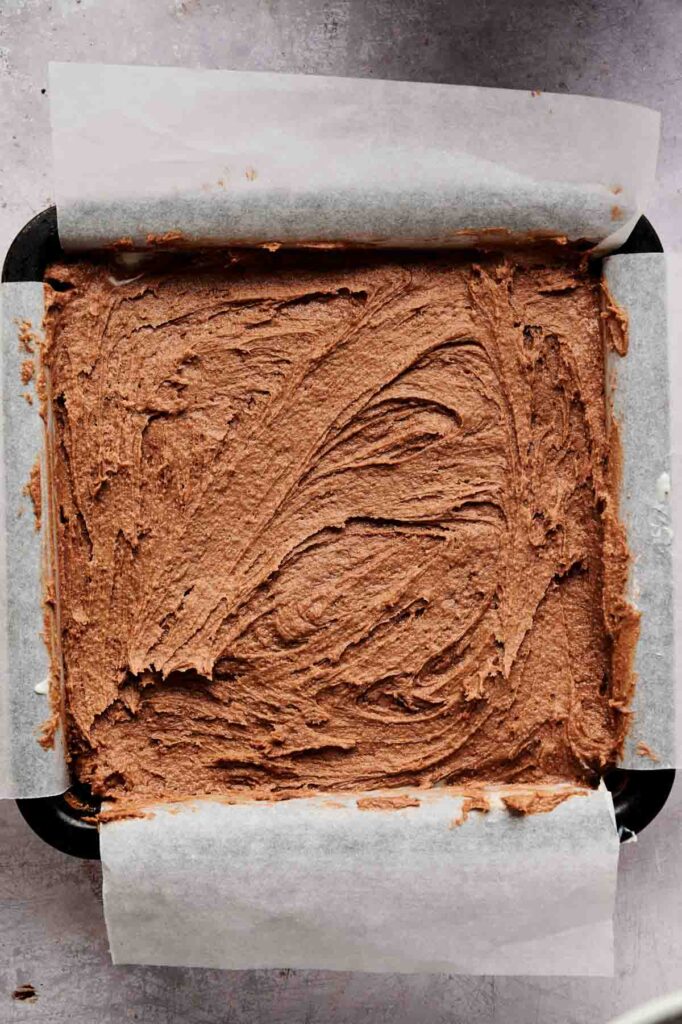A square baking pan lined with parchment paper is filled with rich Air Fryer Cream Cheese Brownie batter, evenly spread and ready to bake.