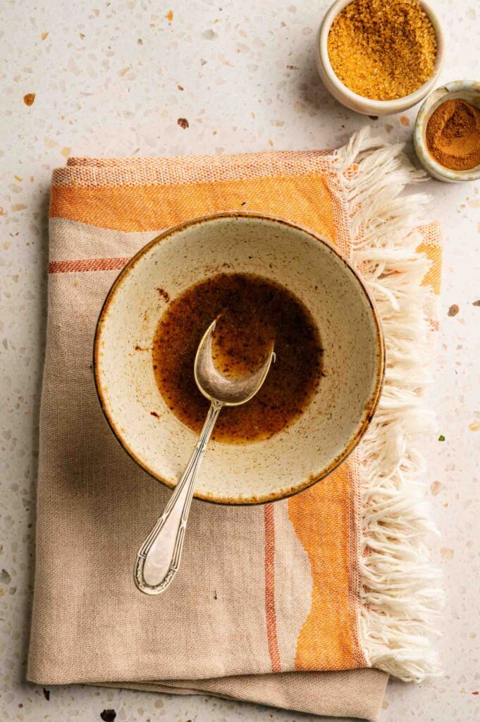 A ceramic bowl with a spoon holds a small amount of brown sauce, perfect for drizzling over grilled peaches, placed on a beige and orange cloth; two small bowls of spices are nearby.