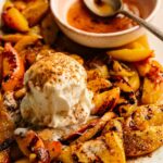A plate of grilled peaches topped with a scoop of vanilla ice cream, sprinkled with cinnamon, served alongside a small bowl of sauce and a spoon.