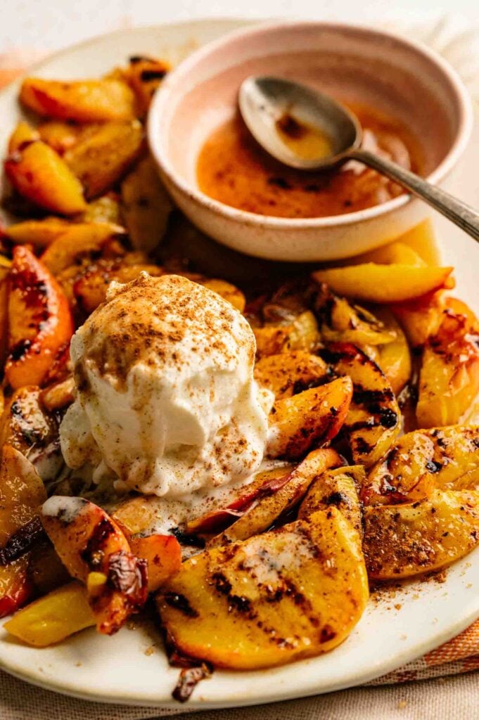 A plate of grilled peaches topped with a scoop of vanilla ice cream, sprinkled with cinnamon, served alongside a small bowl of sauce and a spoon.