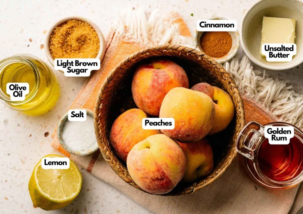 A basket of peaches ready for Grilled Peaches, surrounded by light brown sugar, cinnamon, unsalted butter, golden rum, lemon, salt, and olive oil, all labeled on a white surface.