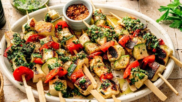 Grilled vegetable and halloumi skewers with red bell pepper, zucchini, and herbs, served on a white platter with a small bowl of crushed red pepper flakes.