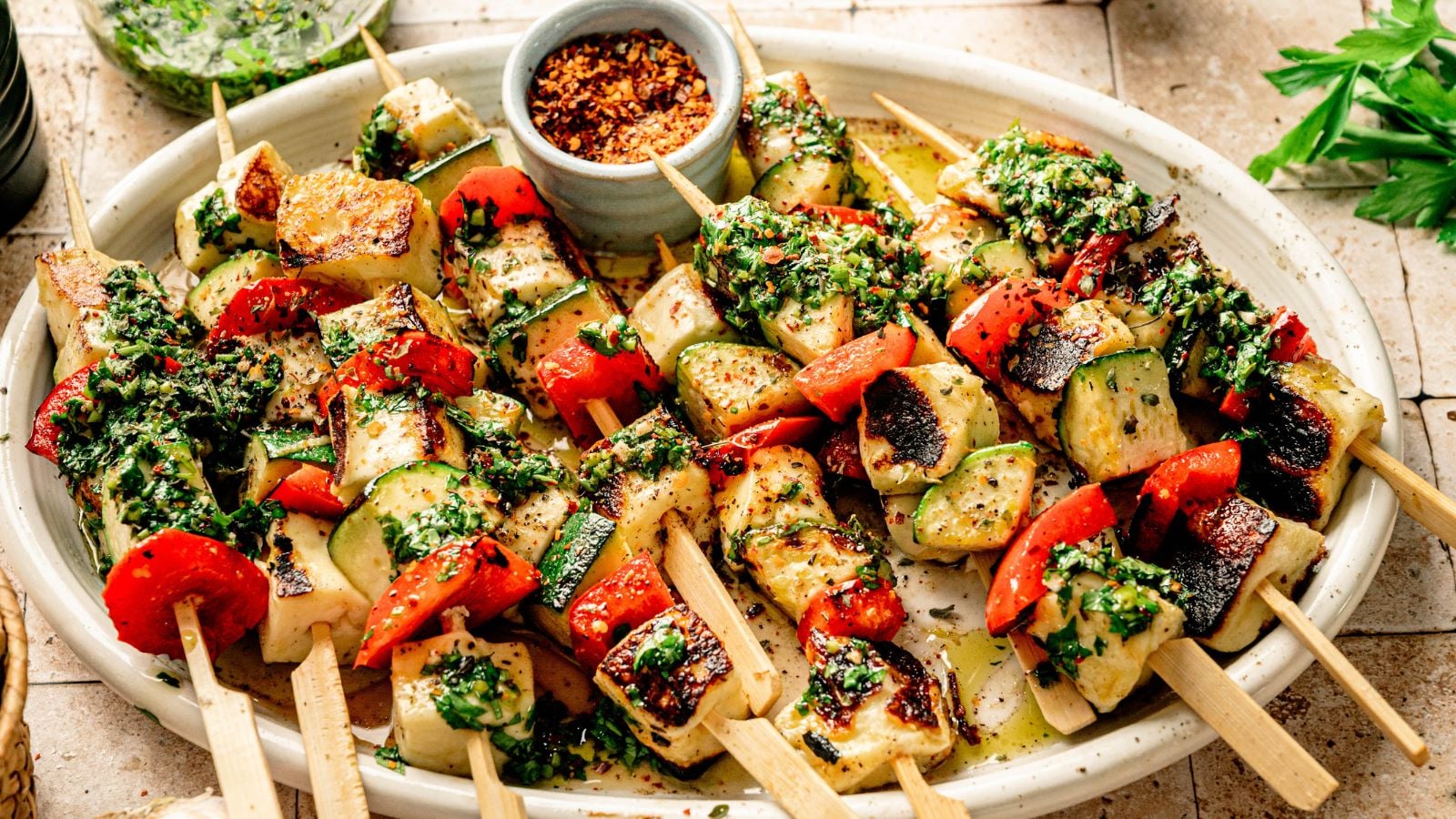 Grilled vegetable and halloumi skewers with red bell pepper, zucchini, and herbs, served on a white platter with a small bowl of crushed red pepper flakes.