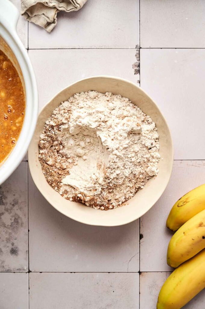 A bowl of flour mixture sits beside a bunch of bananas and a bowl of batter on a tiled kitchen surface, ready for making delicious Air Fryer Pumpkin Banana Bread.