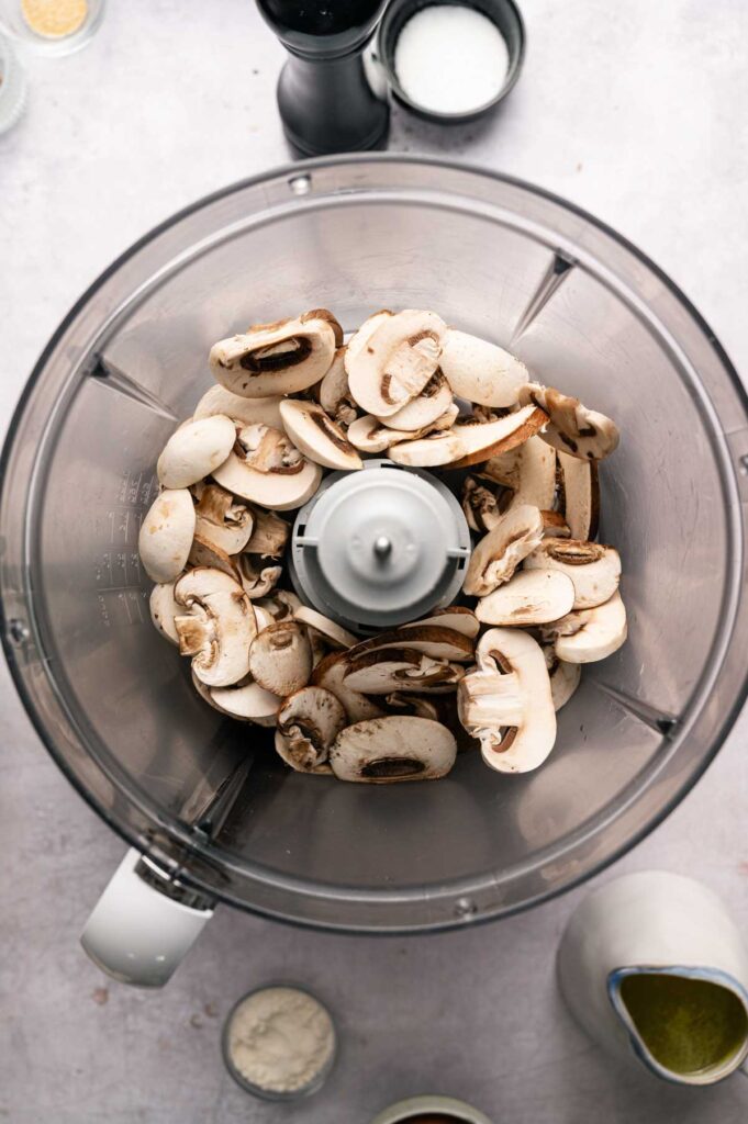 Sliced mushrooms in a food processor bowl, viewed from above, with various bowls and containers of ingredients—perfect for making Air Fryer Vegetarian Meatballs—on the surrounding countertop.