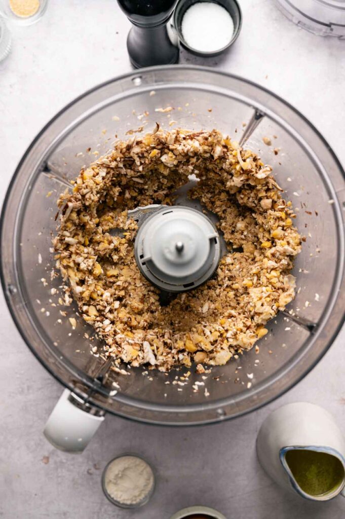 A food processor containing chickpeas and mushrooms sit on a countertop, surrounded by small bowls with various seasonings and a jug of oil—perfect for preparing Air Fryer Vegetarian Meatballs.