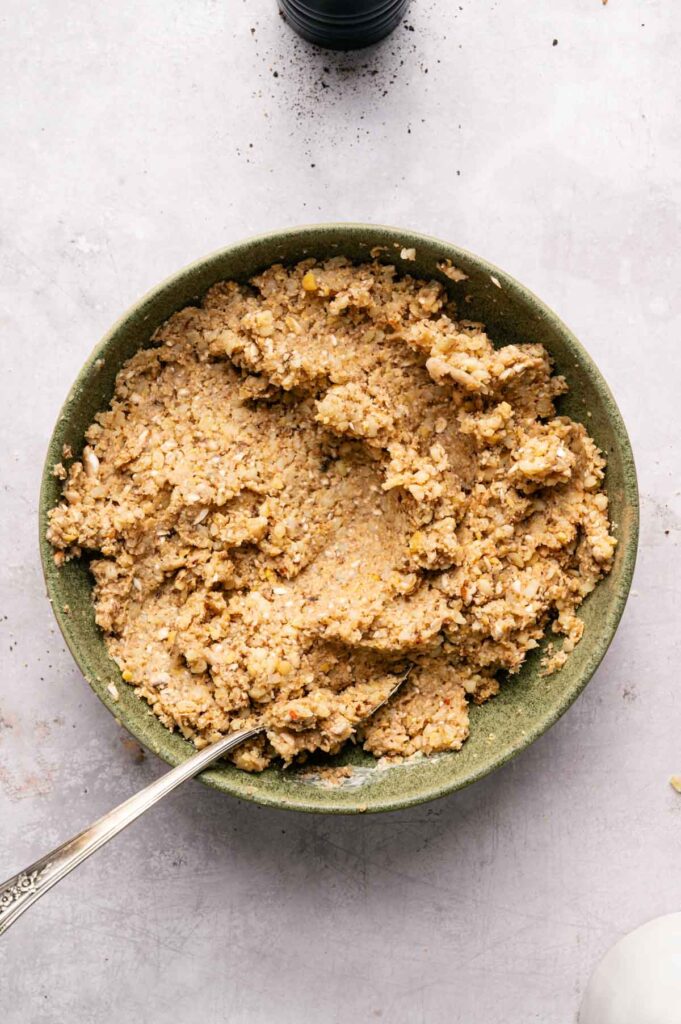 A bowl filled with coarse, beige chickpea mixture for Air Fryer Vegetarian Meatballs sits with a spoon resting inside, placed on a light-colored surface.