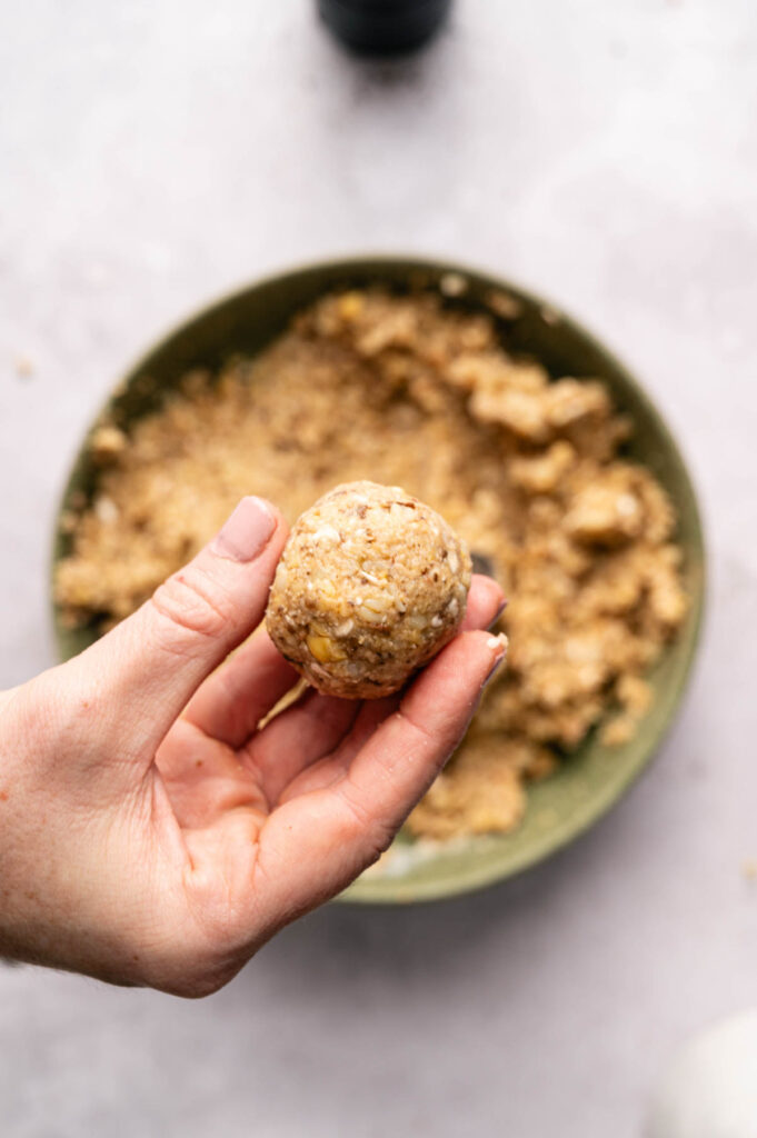 A hand holds a round ball of meatball above a bowl filled with more vegetable mixture on a light countertop, ready to become delicious Air Fryer Vegetarian Meatballs.