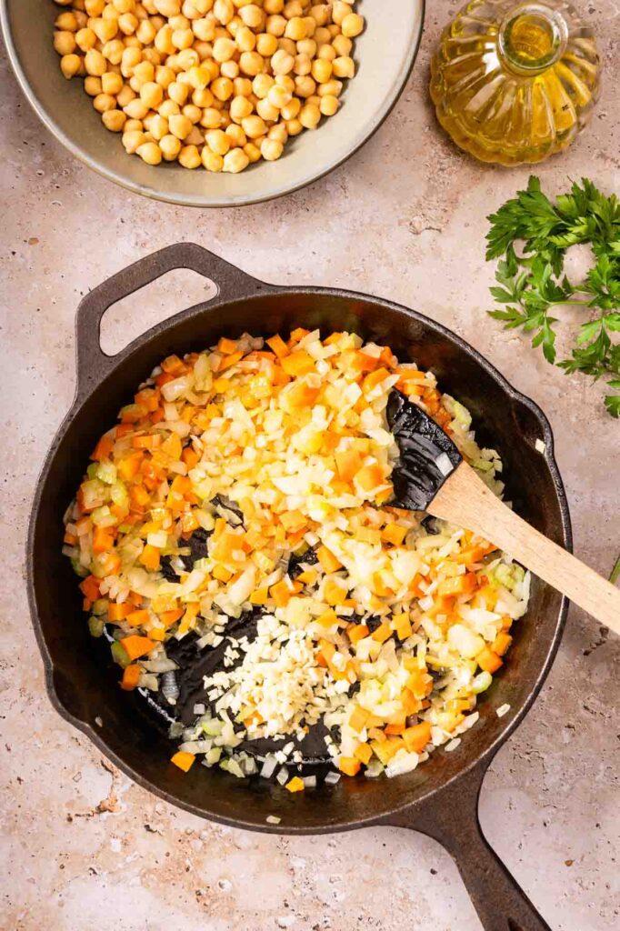 A cast iron skillet with chopped onions, carrots, and garlic being sautéed with a wooden spoon; chickpeas, olive oil, and parsley nearby—perfect ingredients to prep for an Air Fryer Vegetarian Meatloaf.