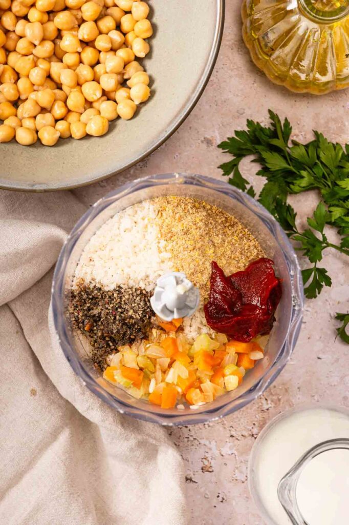 A food processor bowl with chopped vegetables, tomato paste, spices, breadcrumbs, and milk, beside a bowl of chickpeas and fresh parsley—perfect ingredients for preparing Air Fryer Vegetarian Meatloaf.
