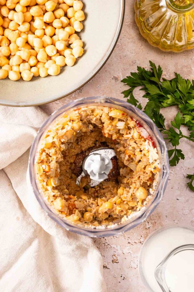 Overhead view of chickpeas mixture in a food processor, surrounded by a plate of chickpeas, parsley, olive oil, and a glass of liquid—perfect ingredients for making Air Fryer Vegetarian Meatloaf on a light countertop.