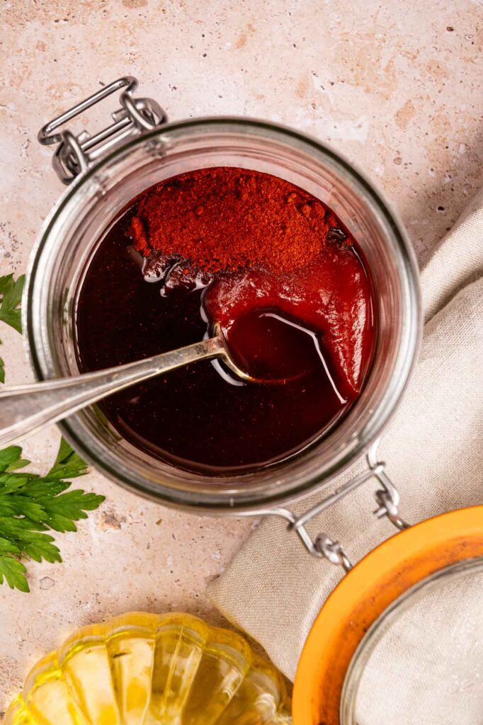 An open glass jar filled with a dark red sauce and a spoon, with spices on top—perfect for drizzling over your Air Fryer Vegetarian Meatloaf. Parsley and a bottle of oil are nearby on a beige surface.