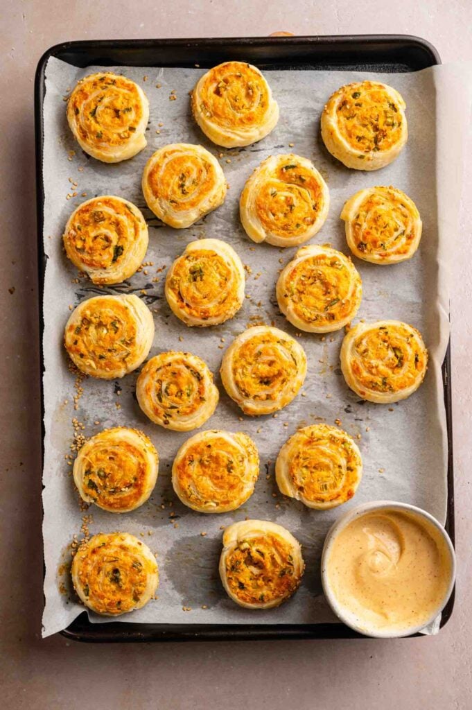 A baking tray lined with parchment paper holds rows of golden Vegetarian Sausage Rolls pinwheels with herbs, alongside a small bowl of dipping sauce.
