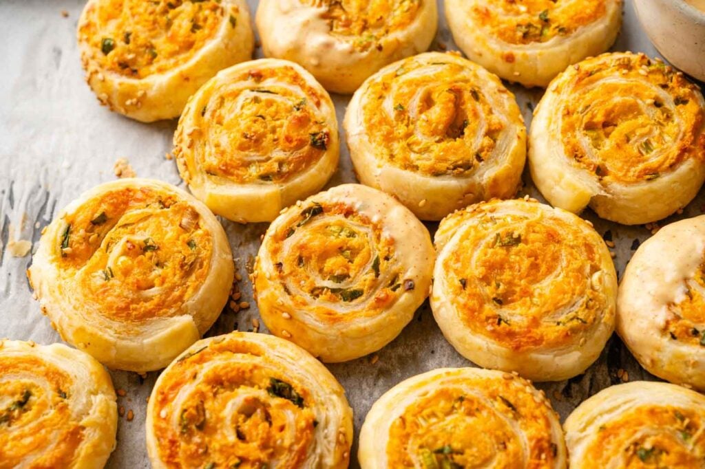 Golden-brown Vegetarian Sausage Rolls and cheese and herb puff pastry pinwheels are arranged on a baking sheet, showing flaky layers and melted cheese.
