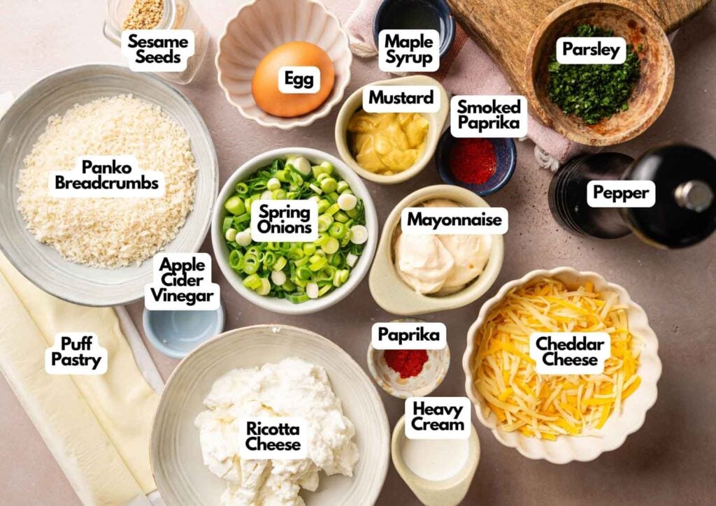 Various labeled ingredients for Vegetarian Sausage Rolls, including egg, sesame seeds, spring onions, puff pastry, ricotta cheese, cheddar, mayonnaise, cream, spices, vinegar, mustard, and parsley are arranged on a table.