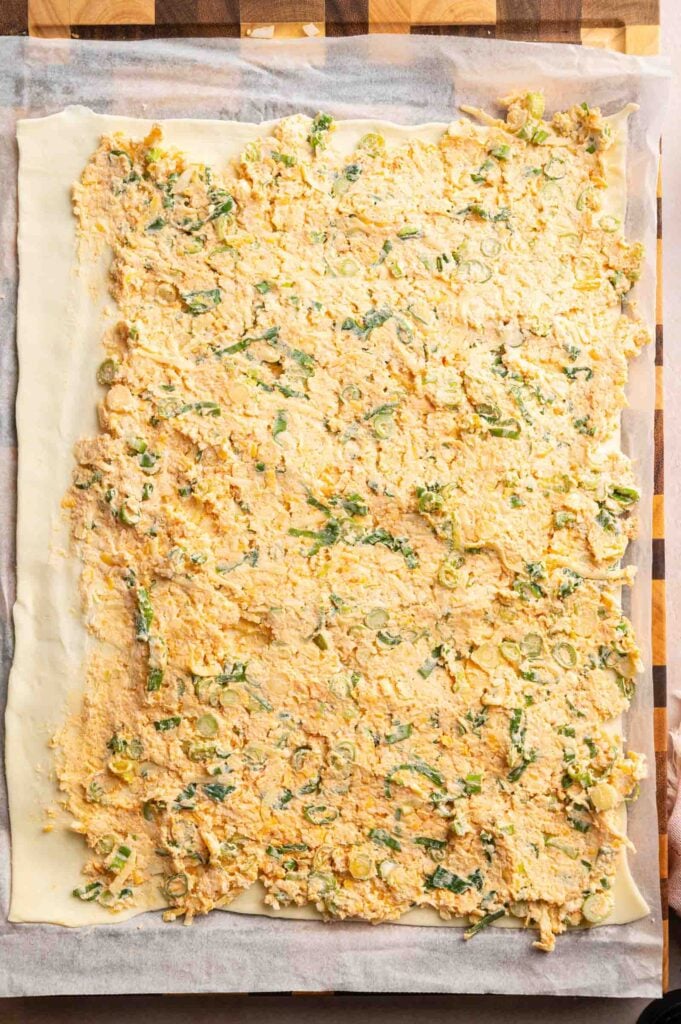 Rectangular sheet of dough on parchment paper topped with an even layer of creamy, vegetarian sausage roll filling with green herbs, ready to be rolled into delicious Vegetarian Sausage Rolls on a wooden surface.