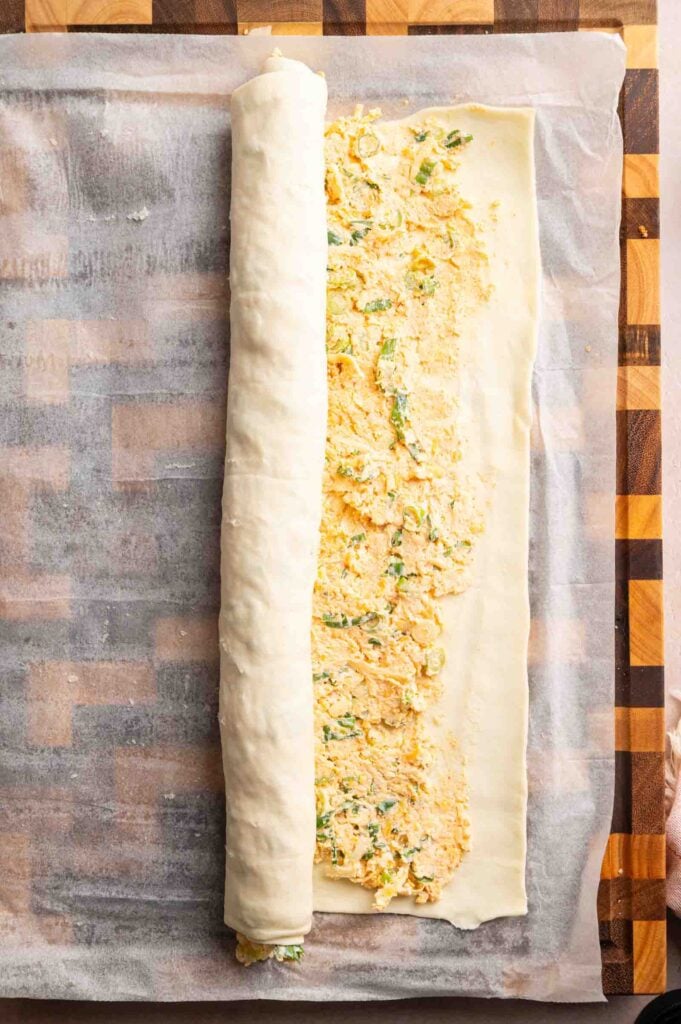 A sheet of pastry dough is being rolled up around a cheese and herb filling, creating delicious Vegetarian Sausage Rolls, on parchment paper atop a wooden surface.
