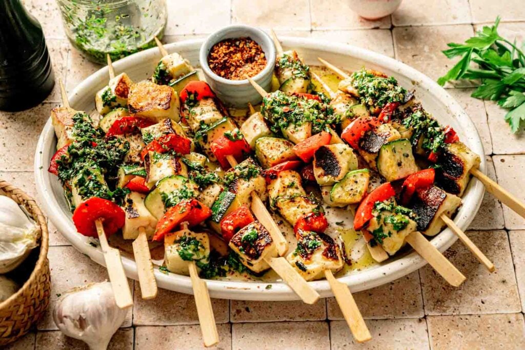 A platter of grilled vegetable, tofu, and halloumi skewers garnished with chopped herbs, served with a small bowl of red pepper flakes and vibrant chimichurri.