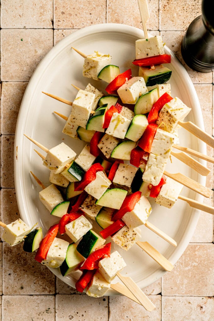 Grilled Halloumi Skewers with Chimichurri feature cubes of halloumi, red bell pepper, and zucchini arranged on a white oval plate atop a tiled surface.