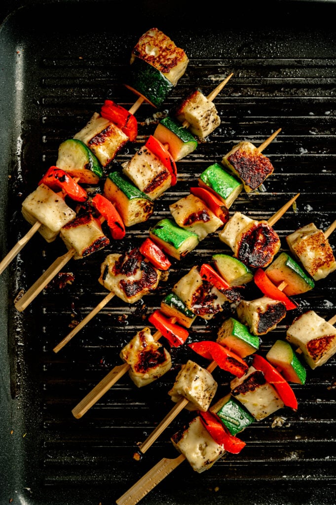 Three Grilled Halloumi Skewers with Chimichurri, featuring zucchini, red bell pepper, and browned cheese, are served on a ridged grill pan for a flavorful twist.