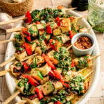 Grilled Halloumi Skewers with Chimichurri, paired with vibrant vegetables, chopped herbs, and spices, are served on a white platter, accompanied by a jar of chimichurri sauce, chili flakes, garlic cloves, and a pepper grinder.