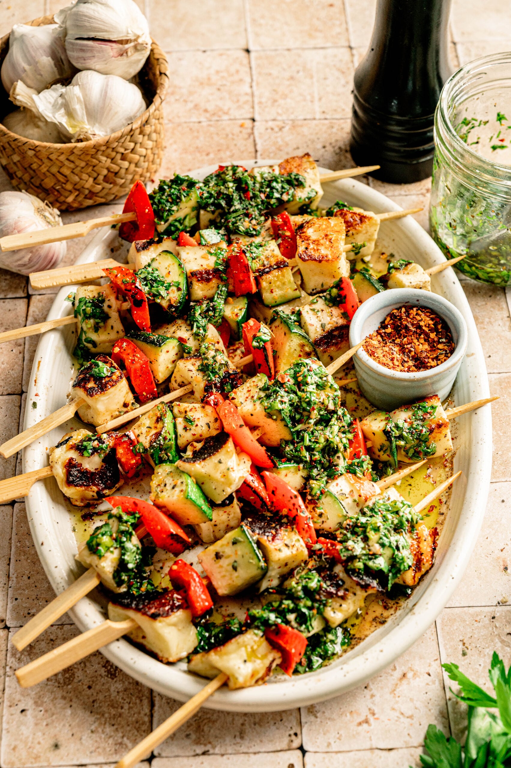 Grilled Halloumi Skewers with Chimichurri, paired with vibrant vegetables, chopped herbs, and spices, are served on a white platter, accompanied by a jar of chimichurri sauce, chili flakes, garlic cloves, and a pepper grinder.