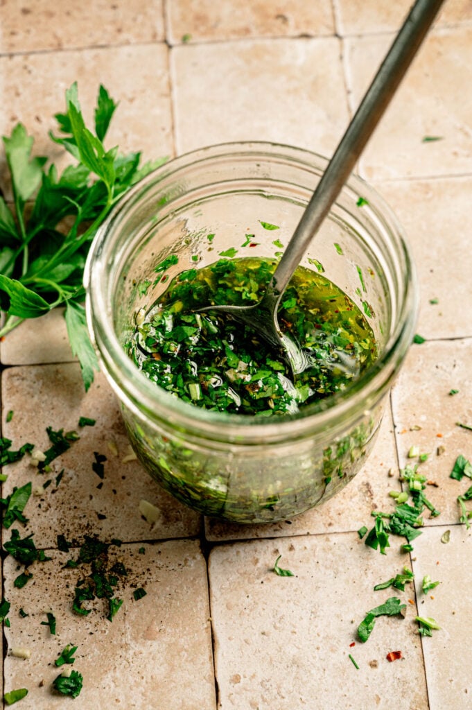 A glass jar filled with chopped herbs and oil mixture for Grilled Halloumi Skewers with Chimichurri, with a spoon inside, sits on a tiled surface next to fresh herbs.