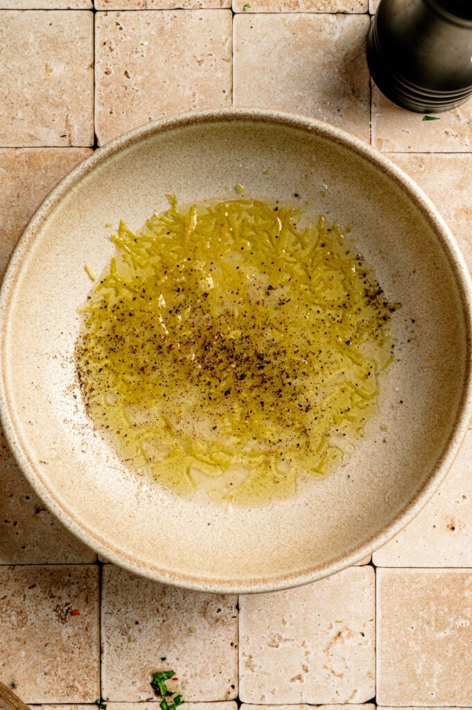 A beige bowl with lemon zest, olive oil, salt, and pepper sits on a tiled surface—perfect for pairing with Grilled Halloumi Skewers with Chimichurri.