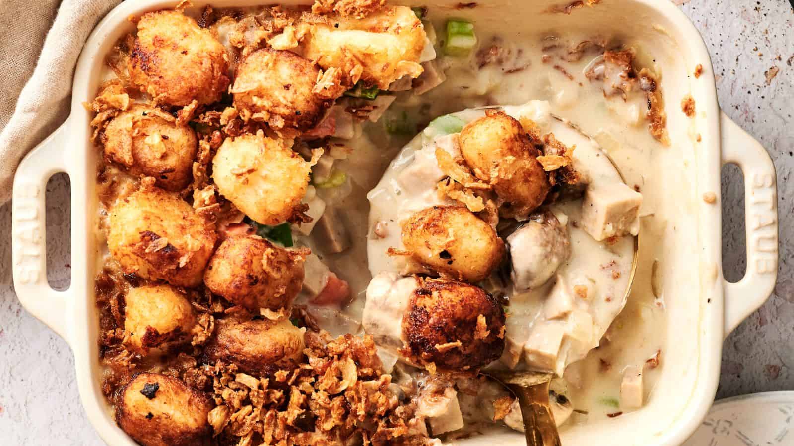 A baked dish featuring a creamy chicken casserole topped with golden brown tater tots and crispy onions in a white oven-safe dish. A portion is scooped out, revealing the creamy filling.